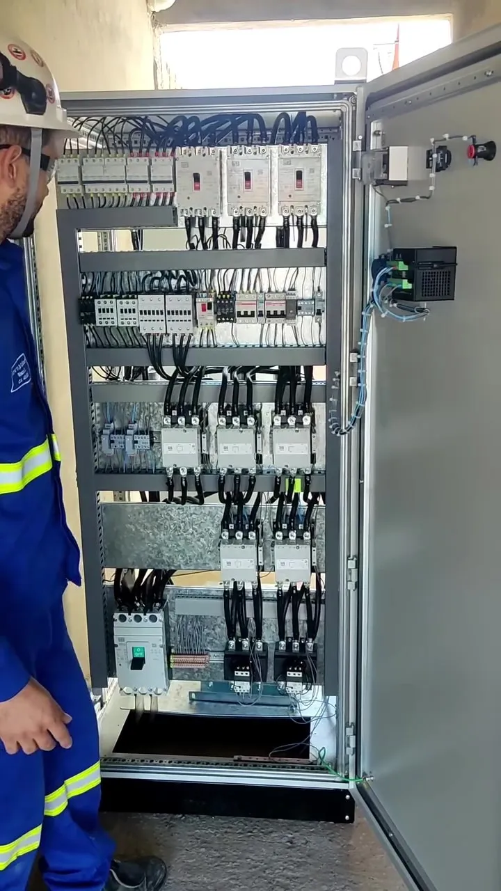 Eletricista com uniforme e capacete de segurança observando um centro de controle de motores aberto, com disjuntores, contatores e cabeamento organizado.