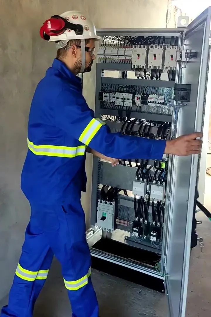 Eletricista da Elétrica Oeste com capacete, protetor auricular, óculos de proteção e macacão com faixa refletiva, abrindo a porta de um painel de comando industrial para intervenção.