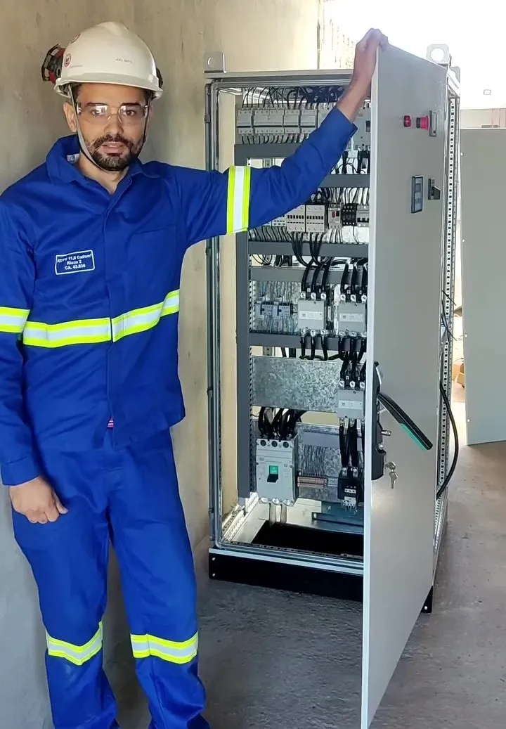 Eletricista com uniforme e EPI ao lado de um painel elétrico aberto e cabeado, com disjuntores e contatores organizados, e outro quadro fechado ao fundo.