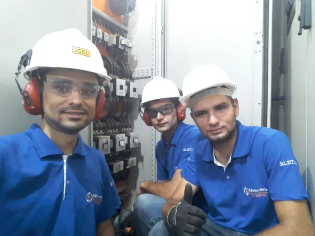 Três eletricistas da Elétrica Oeste com capacete, óculos e protetor auricular, diante de um painel de comando aberto em sala elétrica.
