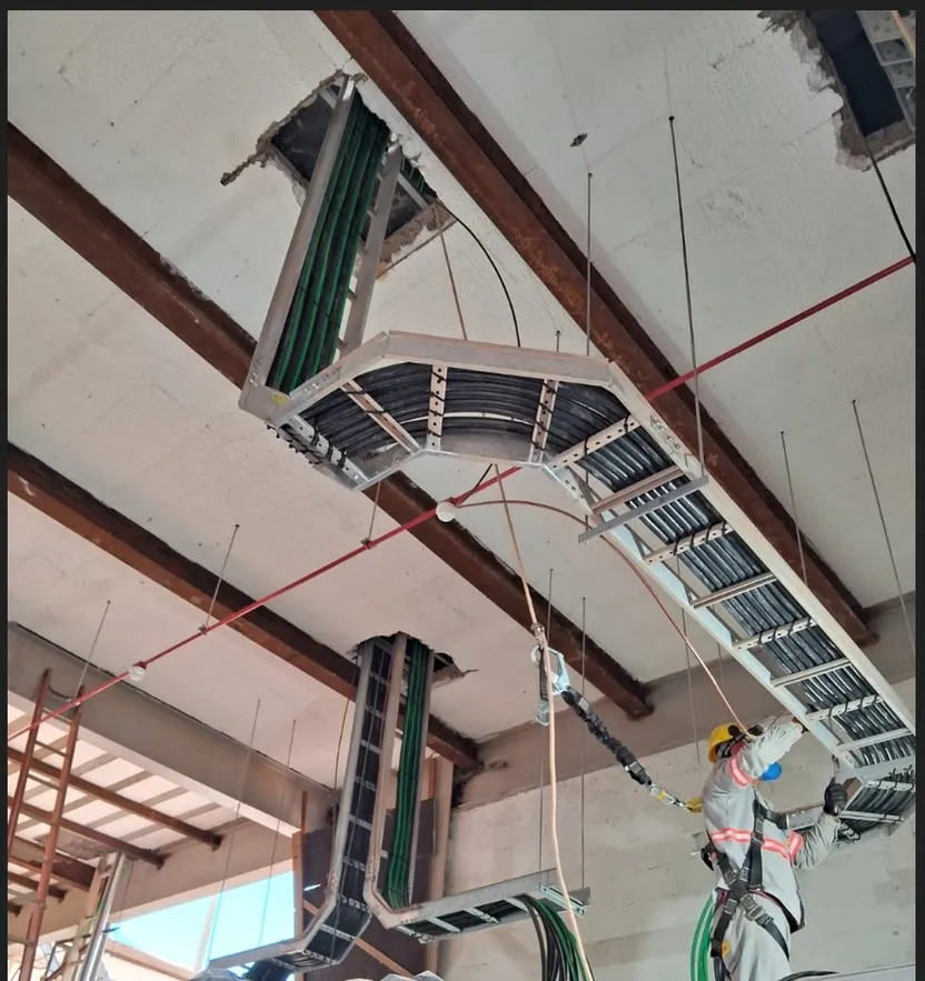 Eletricista com cinto de segurança instalando um trecho curvo de leito de cabos, suspenso sob laje de concreto, com cabos organizados passando por duas aberturas estruturais.