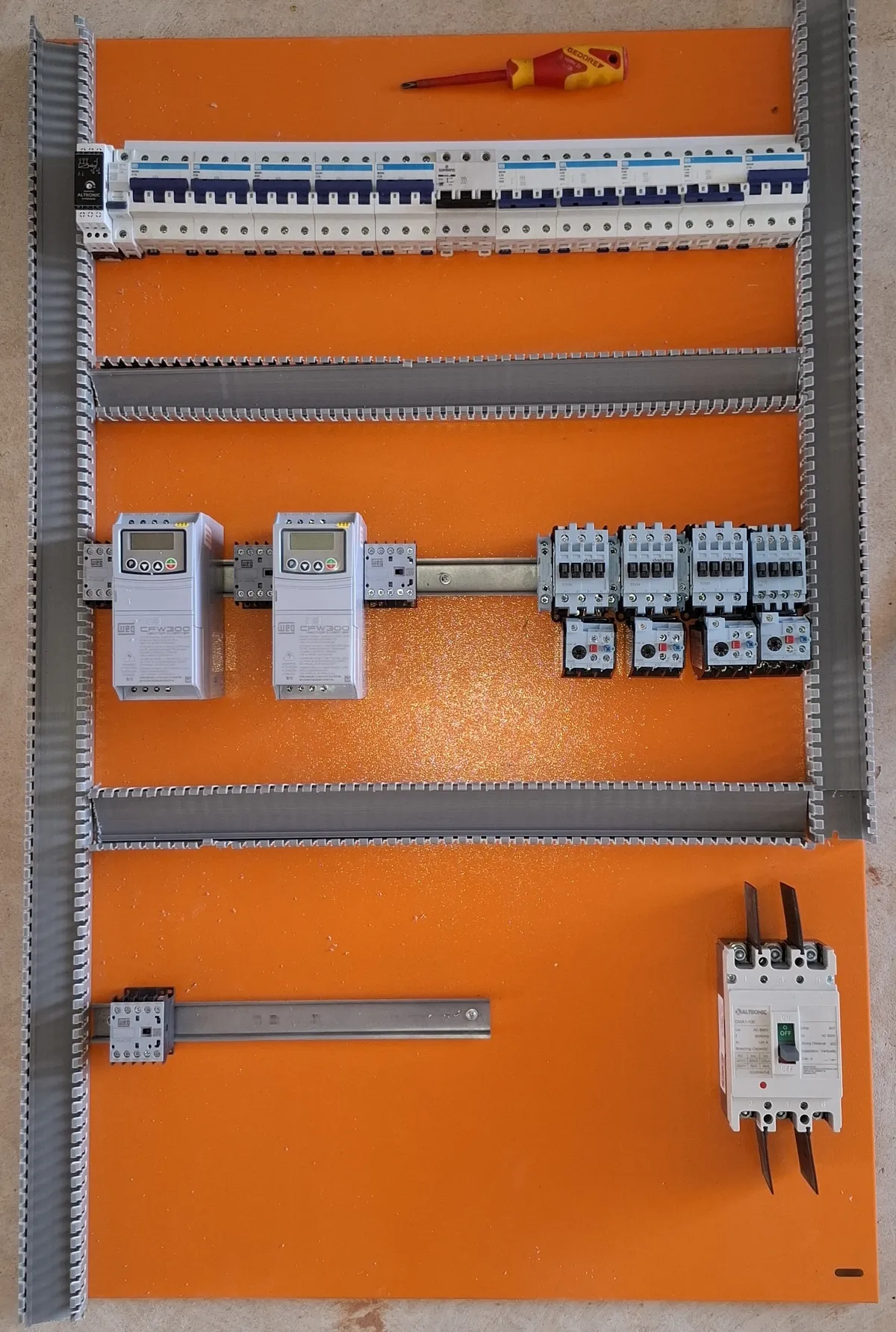 Placa de montagem de painel elétrico apoiada no piso, com uma régua de disjuntores, dois inversores de frequência WEG CFW300 e quatro contatores Altronic com relés térmicos fixados no trilho, ainda sem fiação.