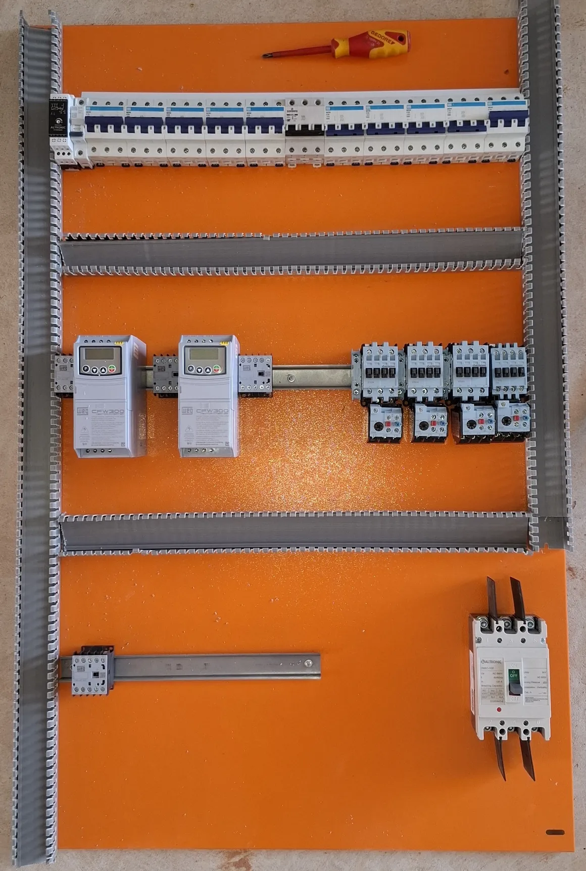 Placa de montagem de painel elétrico apoiada no piso, com uma régua de disjuntores, dois inversores de frequência WEG CFW300 e quatro contatores Altronic com relés térmicos fixados no trilho, ainda sem fiação.