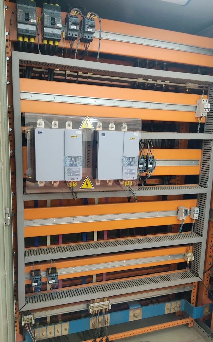 Painel elétrico aberto com barramentos sobre placa laranja, disjuntores-motor, contatores e dois soft-starters WEG SSW-07 montados lado a lado.