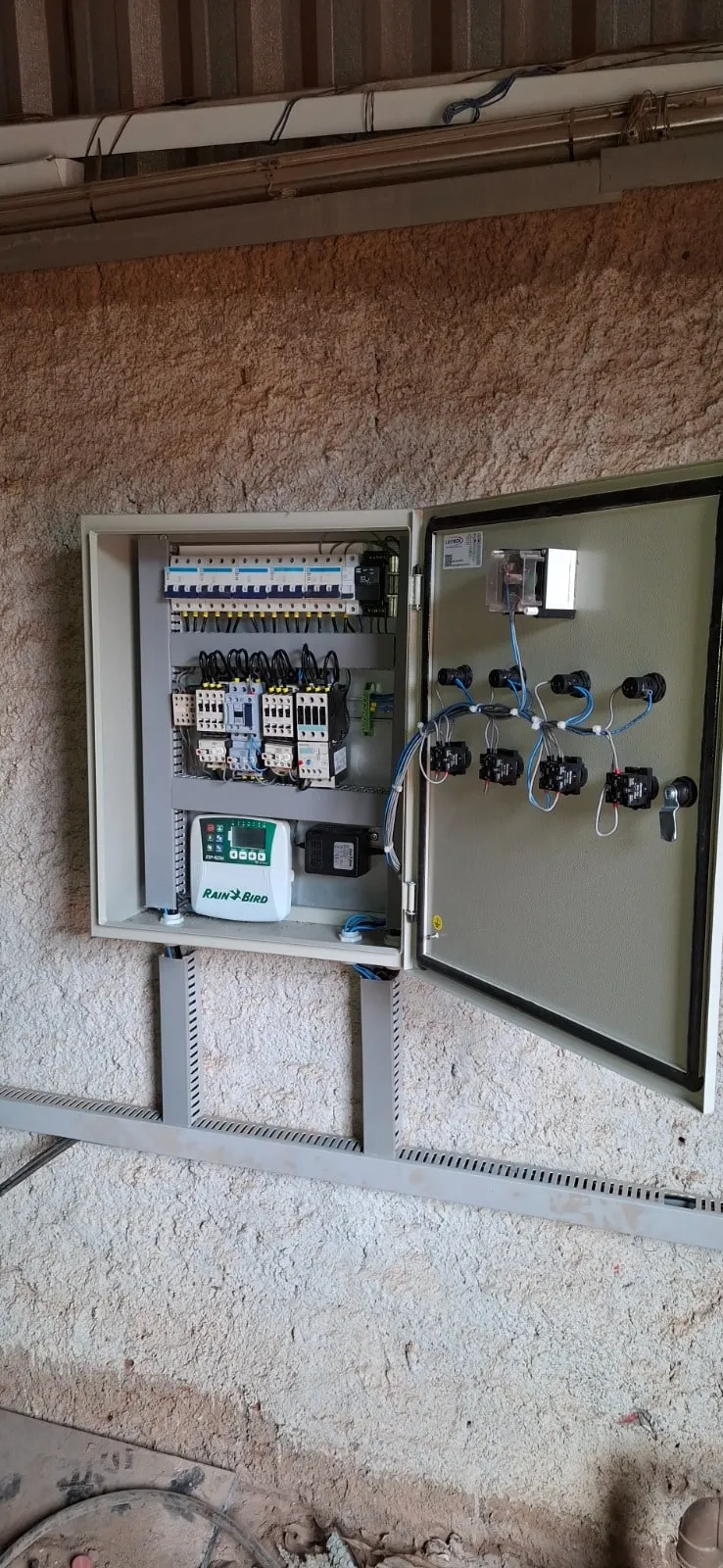 Quadro de comando de irrigação aberto, montado em parede externa, com disjuntores, contatores e um controlador Rain Bird ESP-RZXe.