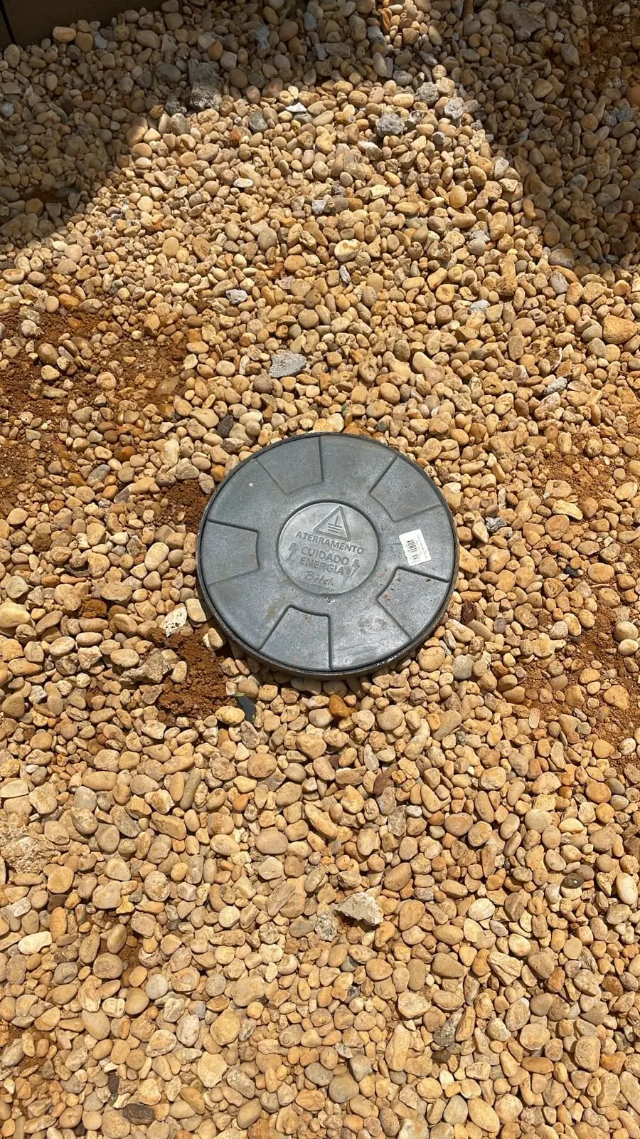 Caixa de inspeção de aterramento fechada, instalada ao nível do solo sobre leito de brita, com a tampa identificando aterramento.